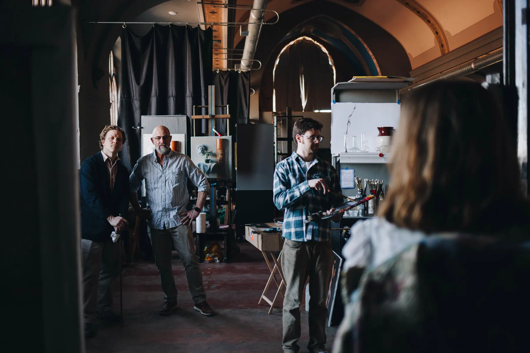 A group of three men stand in an art studio with high ceilings and arched windows. One man holds a palette, while another leans on a cane. A fourth person with long hair is in the foreground, facing them. The room is filled with art supplies.