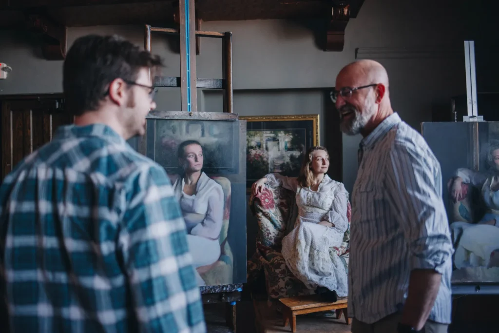 Three people in an art studio, two men discussing paintings on easels. A seated woman in the background is the subject of the portraits. The room has a classic, artistic ambiance with wooden beams and framed art.