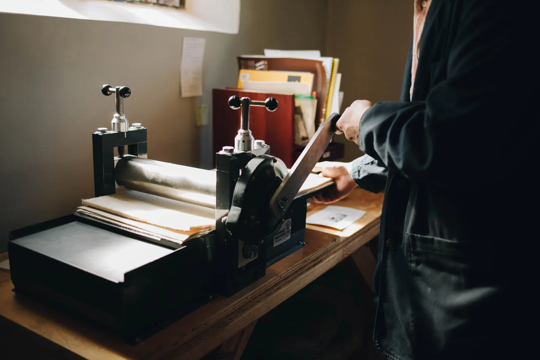 A person operating a printing press with two large handles, pressing paper or material between rollers. The machine is on a wooden table, and there are shelves with papers and folders in the background. Natural light enters from a nearby window.