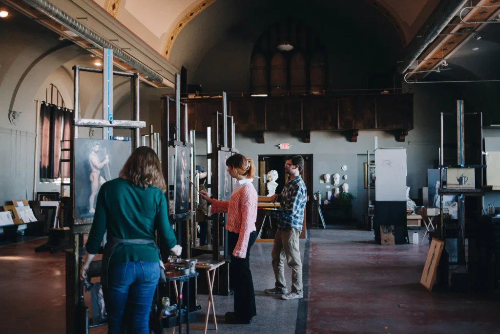 People in an art studio are painting on canvases. They are surrounded by various art supplies and sculptures. The studio has tall windows and arched ceilings, creating a spacious atmosphere.