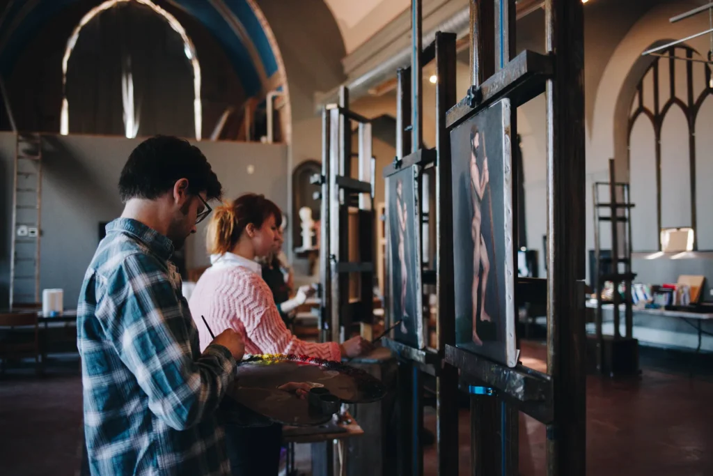 Artists concentrating on their easels in a well-lit studio with high arched windows and paintings displayed on wooden stands. The space has an artistic and creative ambiance.