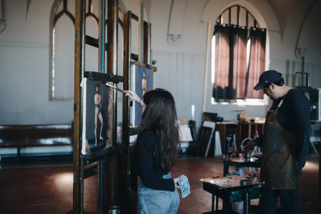 A woman with long hair paints a figure on a canvas in an art studio. A man wearing a cap and apron stands nearby observing. The room has large arches and a warm atmosphere, with light streaming through partially drawn curtains.