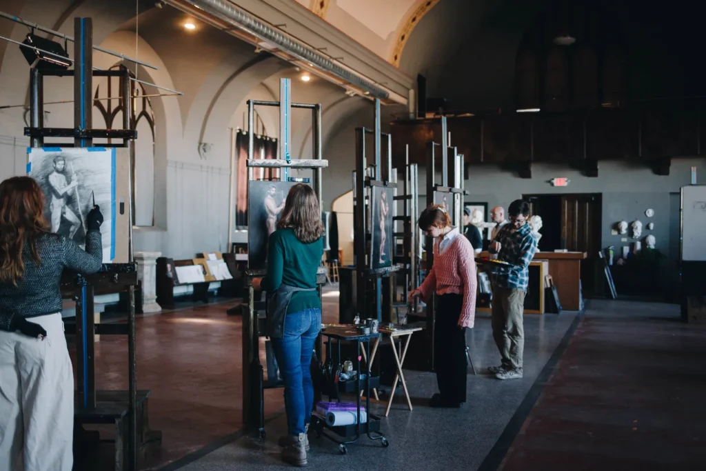 A group of artists stands at easels in a spacious, well-lit studio with arched ceilings, painting or drawing. The room has a classic design and various art supplies are scattered around. Each person is focused on their canvas.