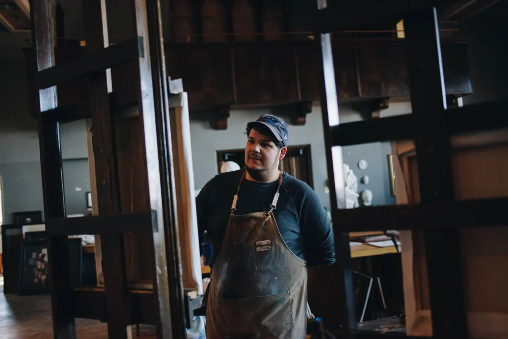 A person wearing a cap and an apron stands among large wooden frames in a workshop. The background features woodworking tools and various pieces of artwork. The person appears focused and engaged in their work environment.