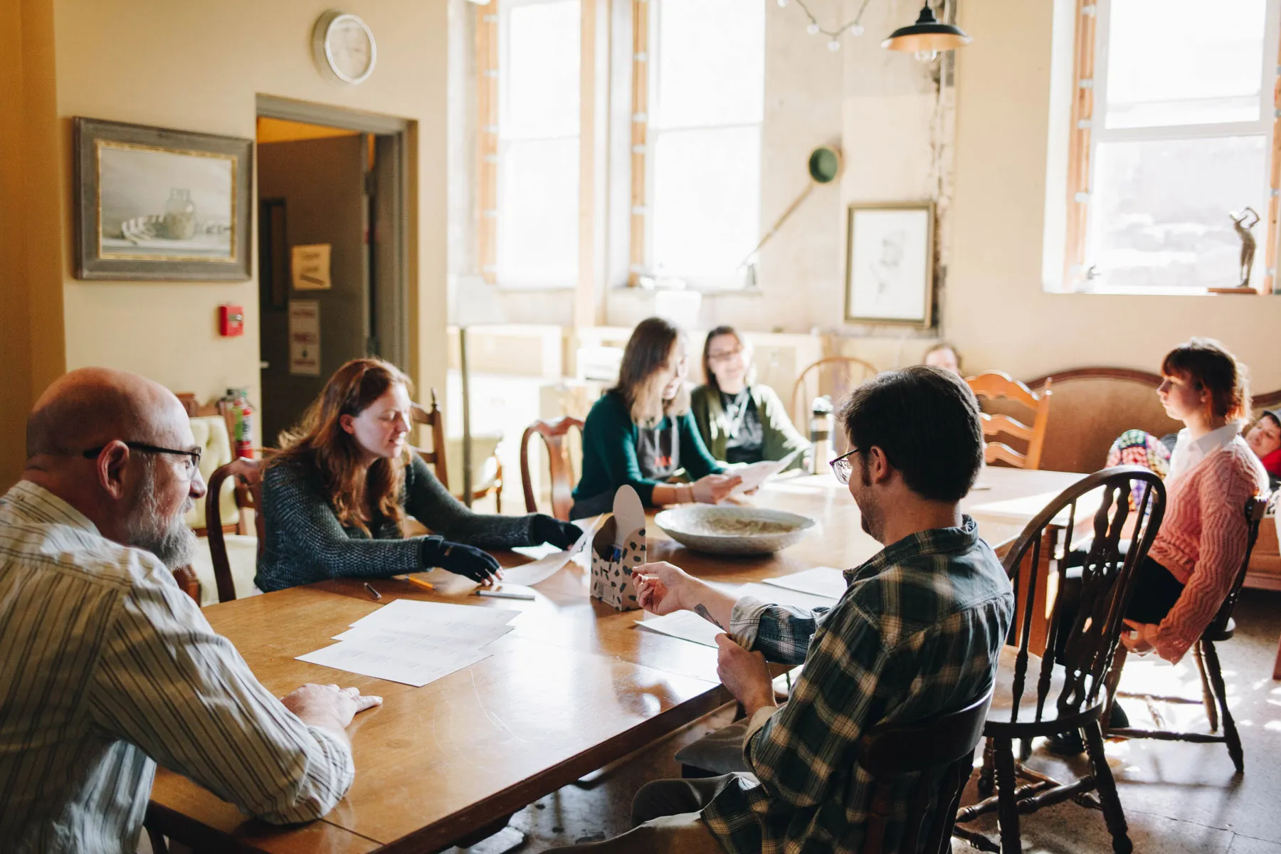 A group of people sitting around a large wooden table in a sunlit room with papers and a coffee cup. The room has framed art, large windows, and a mix of wooden chairs. The atmosphere appears casual and collaborative.