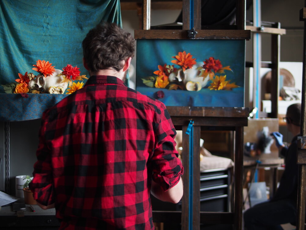 A person in a red and black checkered shirt stands in front of an easel painting a still life. The still life on the easel features colorful flowers and a skull against a blue draped background. Another person is seated nearby.