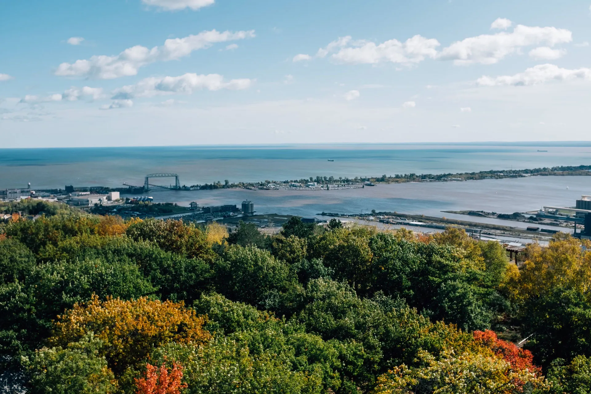 A scenic view of a large body of water under a blue sky with scattered clouds. In the foreground, lush green trees with hints of autumn colors are visible. The distant shoreline features industrial buildings and a prominent structure.